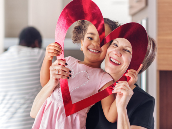 A joyful young girl and a middle-aged woman peek through a large, red heart-shaped cutout, both smiling, with another person visible in the background.