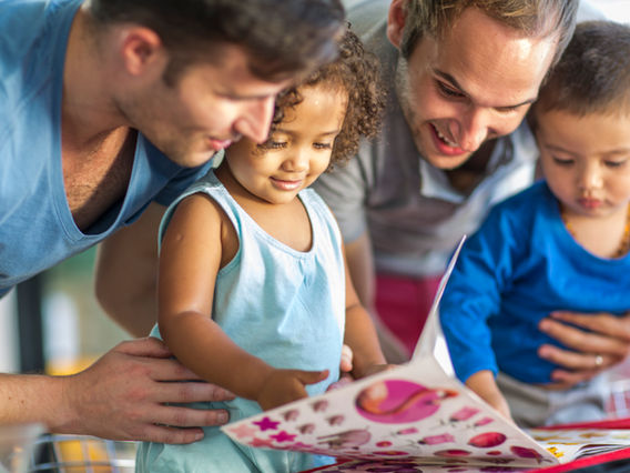 Two men and two young children engaging happily with a picture book. one man is pointing at the book, explaining something to the attentive children. they are seated indoors with natural light.