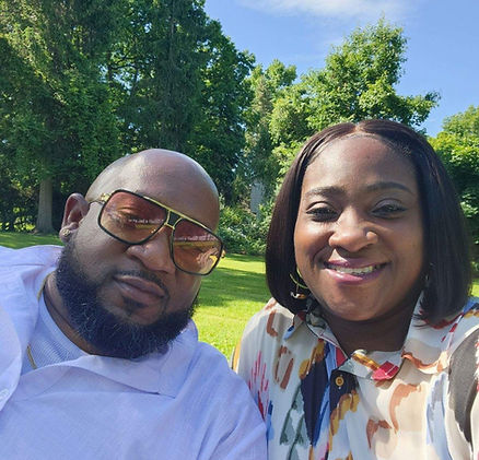 A smiling man and woman posing together in a sunny park setting, both wearing sunglasses, with trees and lush greenery in the background.