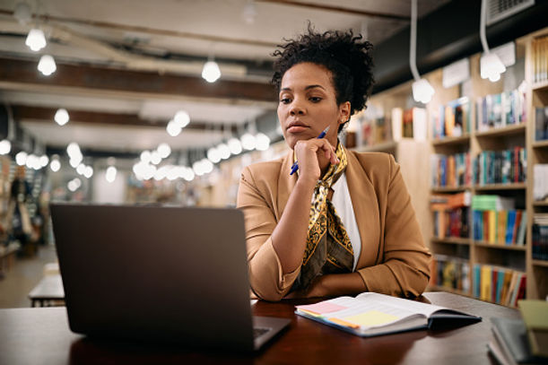 A focused woman sits at a table in a library, using a laptop with books around her. she holds a pen to her chin, deep in thought, surrounded by bookshelves.