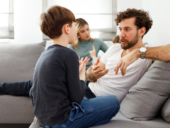 A father and son sit on a couch having a serious conversation, gesturing with their hands, while a woman looks on from the background.