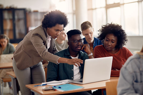 A diverse group of five adult students, including an afro-haired woman instructing, are engaged around a laptop in a bright classroom setting.