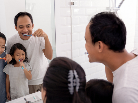 A family engaging in their dental hygiene routine in the bathroom, with a father and his daughter brushing their teeth while smiling at each other in the mirror.