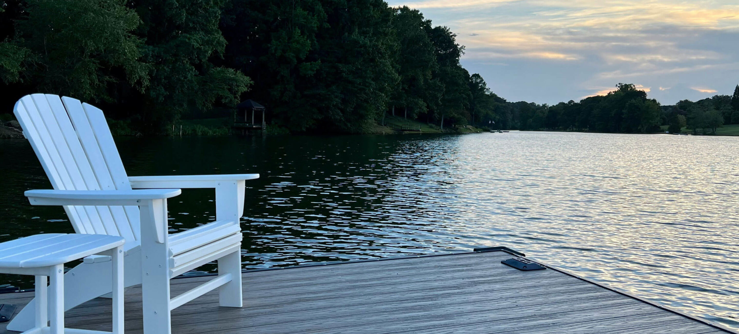 A white adirondack chair sits on a wooden dock, overlooking a tranquil lake with trees lining the far shore under a colorful sunset sky.