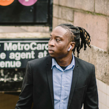 A man with dreadlocks, wearing a blue shirt and earbuds, looks thoughtfully to the side near a subway station entrance.