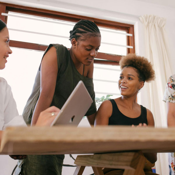 Three women engage in a discussion around a wooden table, one holding a digital tablet. they appear focused and involved in a collaborative work environment.