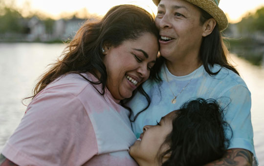 Three people of asian descent joyfully embracing outdoors, with a beautiful sunset over a lake in the background.