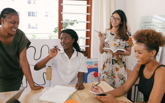 Four women collaborating joyfully around a table in a bright workspace, one writing on a board and the others engaged in discussion with papers and a phone.