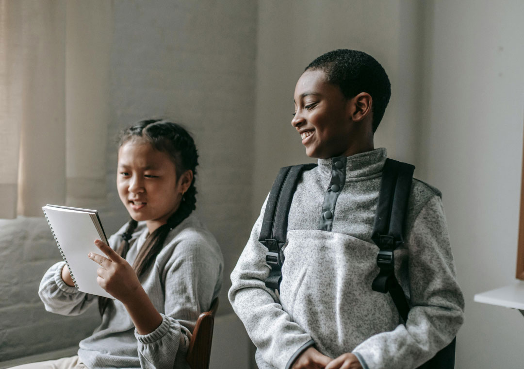 Two students with backpacks sit together; one laughs joyfully while looking at a notebook held by the other, who is speaking and making a facial expression of discomfort or disagreement.