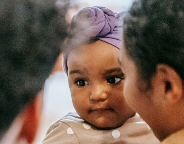 A toddler with a purple headband looks thoughtfully past another child in the foreground, in an indoor, warmly lit setting.