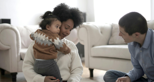A mother sitting on a couch holding her toddler, as a young man sits nearby, looking at the child with interest. all are in a bright living room.