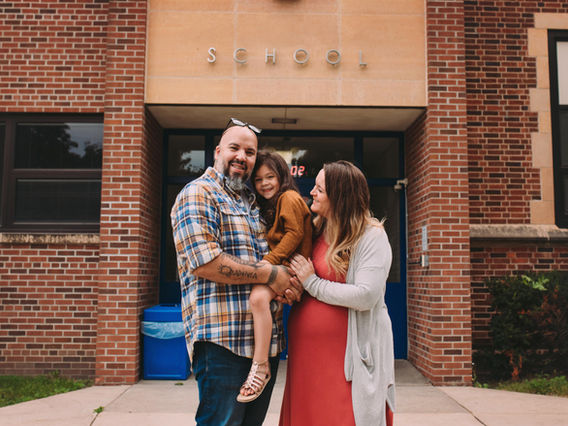 A happy family of three, with a man and a woman holding a young girl, stands smiling in front of a school building. the word 