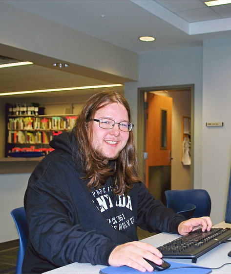 A smiling man with long hair, wearing glasses and a black hoodie, sits at a computer desk in a library with shelves of books in the background.