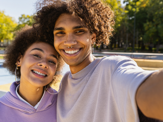 A young, cheerful couple takes a selfie outdoors on a sunny day, both smiling widely at the camera. she wears a purple hoodie, and he wears a gray shirt. trees and a fountain in the background.