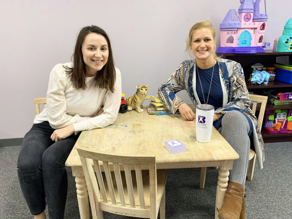 Two women smiling at a small table in a child-friendly room with toys and colorful decorations around, including a castle and toy animals.