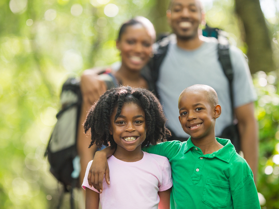 A happy family with two children, smiling in a sunny forest setting, enjoying a day of hiking. the parents are in the background, and the children are in front.
