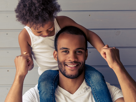 A joyful young father with a beard, wearing a white t-shirt and denim jacket, gives his small daughter a piggyback ride. she, in a white tank top, laughs while holding his hands for support.