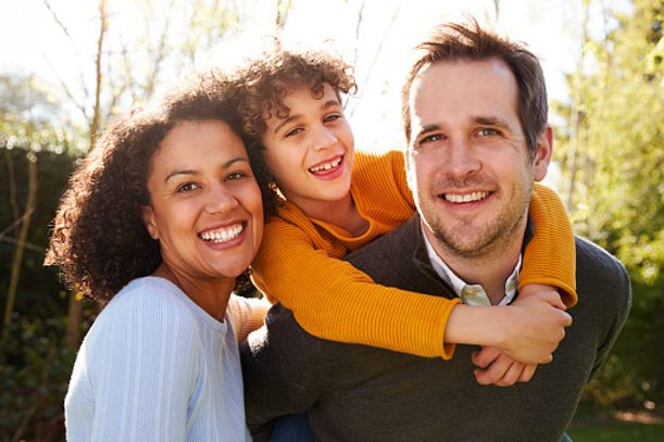 A joyful family outdoors with the father carrying the son on his back and the mother beside them, all smiling broadly. sunlight filters through trees in the background.