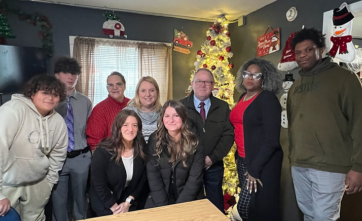 Group of people posing in a room decorated for christmas, with a christmas tree and festive decorations in the background. two people are seated at a table in the front.