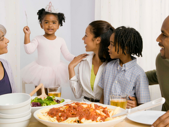 Family sits around a dinner table. They are looking at the little girl who is wearing a tiara.