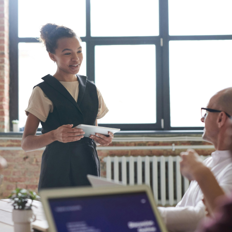 Young woman presenting a document to a colleague in a bright office with large windows.