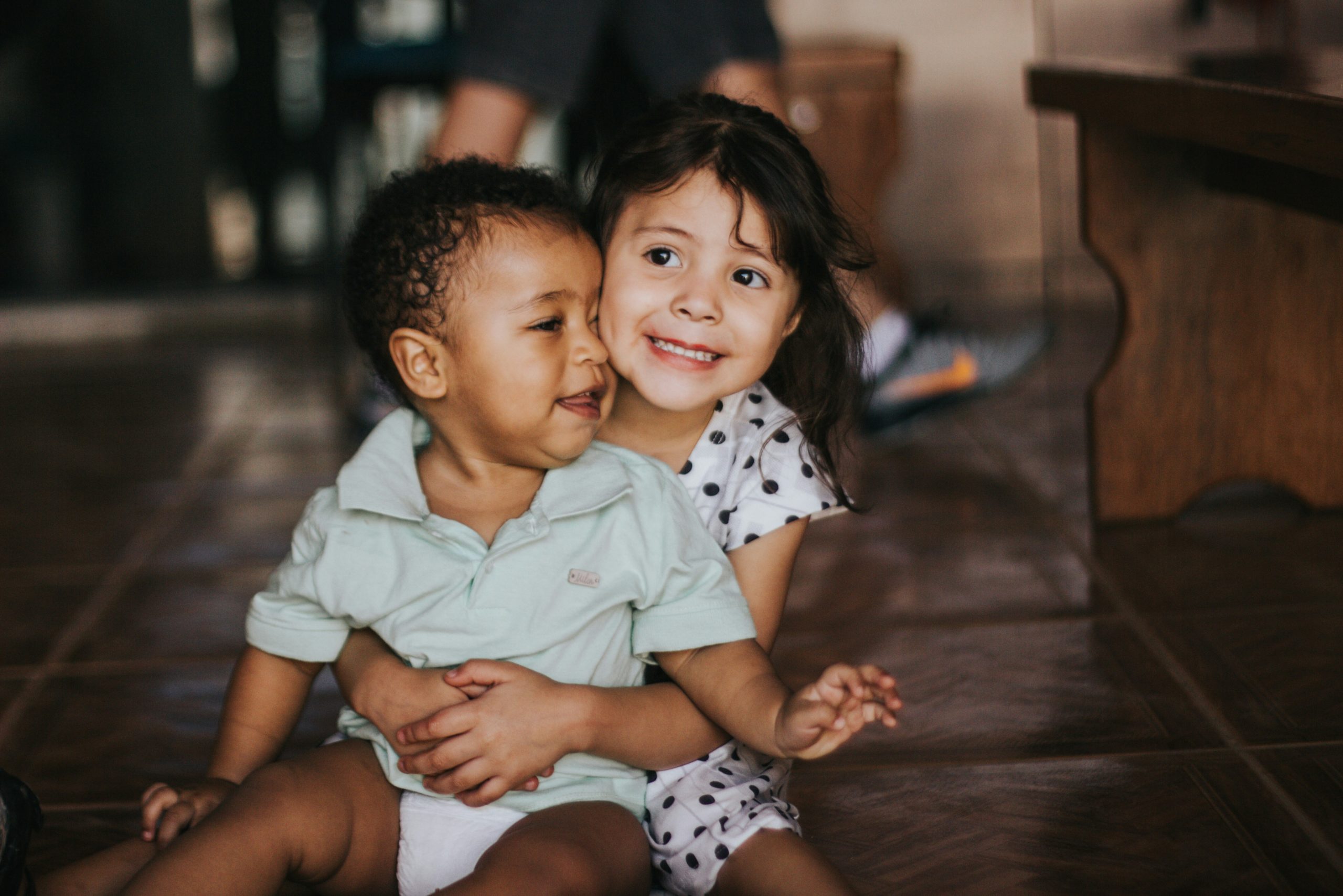 Two young children, a boy and a girl, sit closely together on a tiled floor, smiling joyfully. the boy in a light green polo shirt embraces the girl in a polka dot dress.