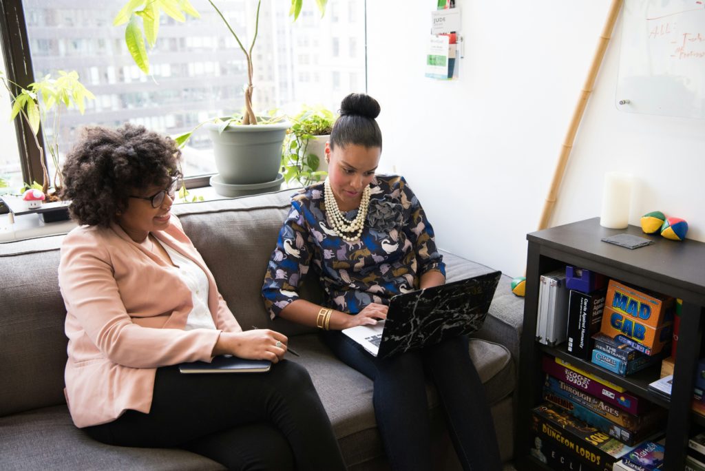 Two women sit on a couch and look at a laptop in an office setting.