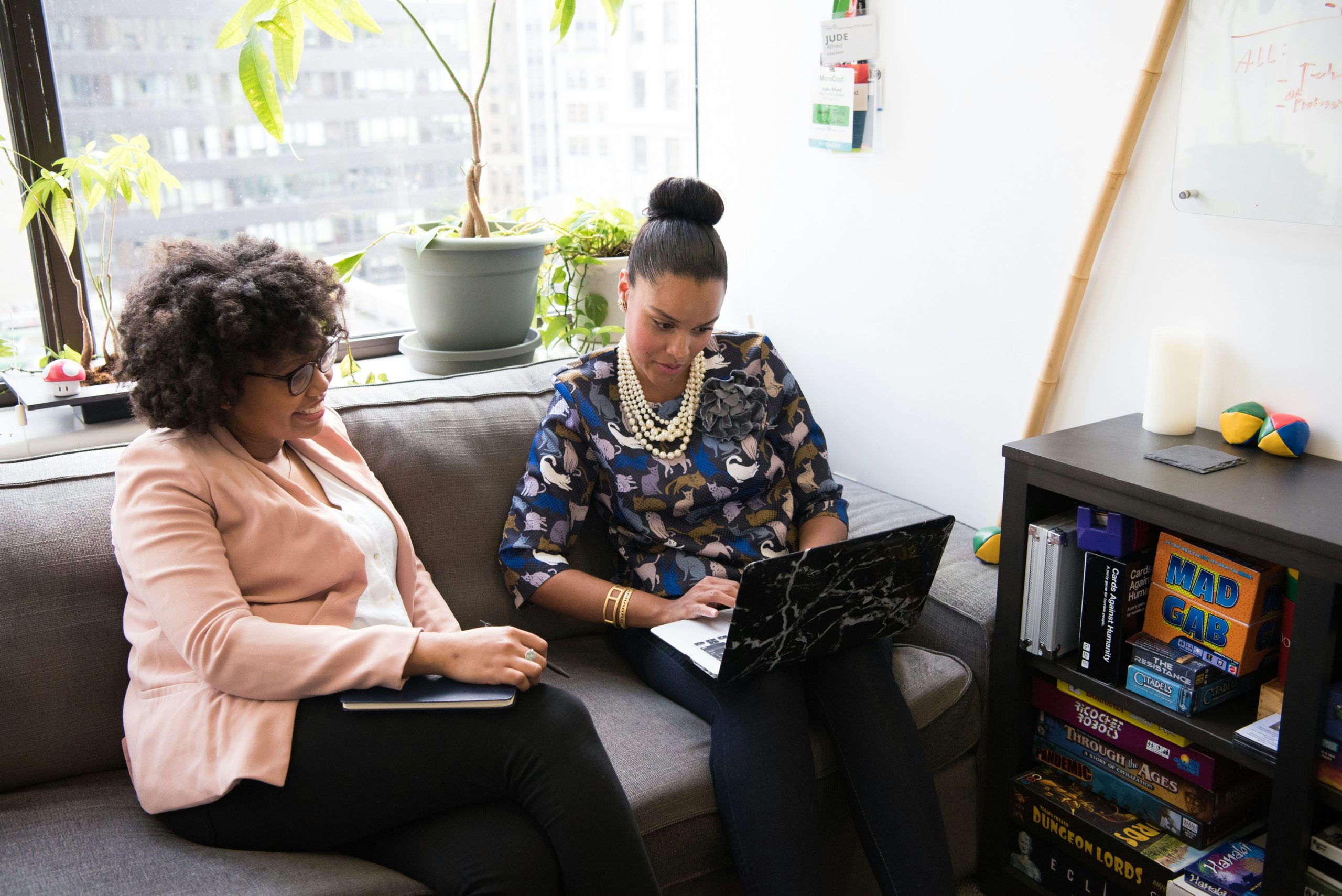 Two women sit on a couch and look at a laptop in an office setting.