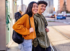 Two joyful young adults, a woman in an orange sweater and a man in a green jacket, walking in a city setting with backpacks, smiling and enjoying a sunny day.