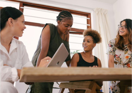 Four women of diverse ethnic backgrounds engaging in a conversation around a wooden table, one holding a digital tablet. they appear cheerful and involved in a collaborative discussion in a bright room.