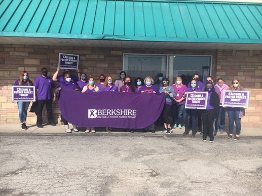 A group of people standing in front of a building, holding a large purple banner reading 