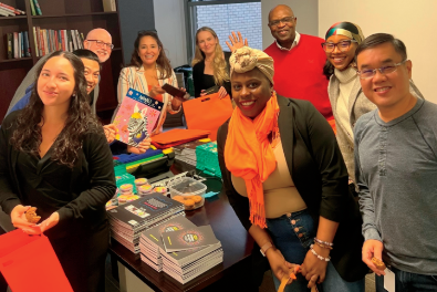 A group of nine people stands together in an office space, smiling at the camera. They are engaged in preparing supplies, with various items like notebooks, bags, cupcakes, and colorful folders on the table in front of them. Shelves with books are visible in the background.