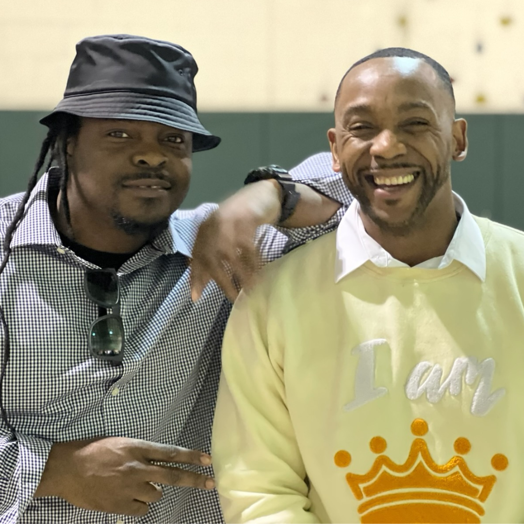 Two men, Tammar and Angelo, posing together indoors, the one on the left wearing a gray hat and sunglasses, the one on the right wearing a yellow sweater, both smiling at the camera.