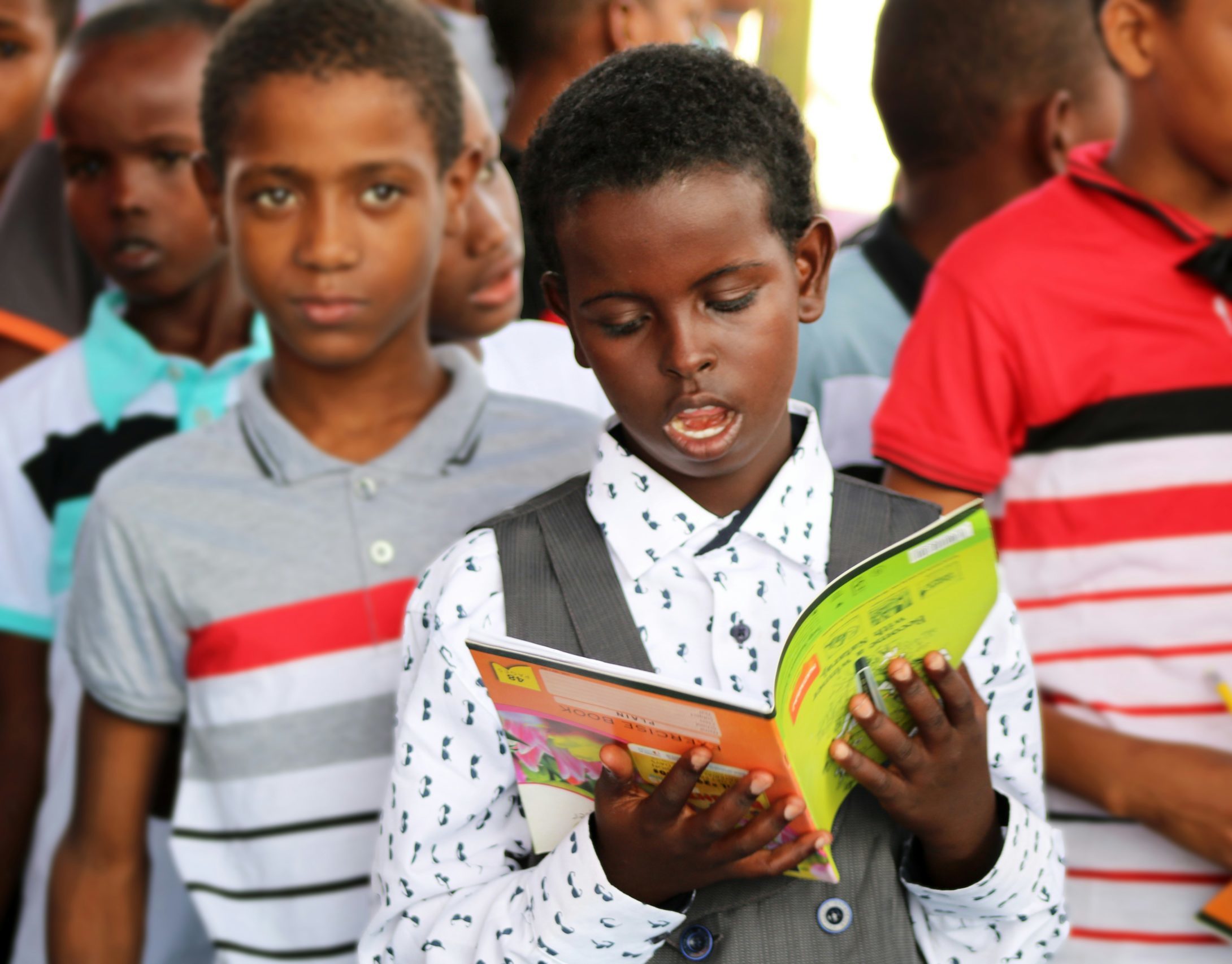 Young boy reads aloud with a group of boys of a similar age behind him.
